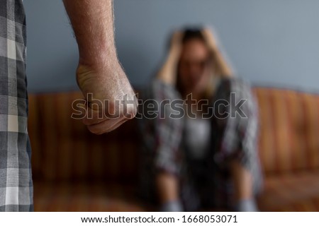 Photo of young woman sitting on sofa at home,focus is on man's fist in the foreground of the image.Home violence concept.Frightened woman and men's fist.Woman is victim of domestic violence and abuse.