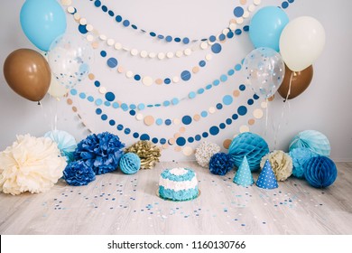 Photo zone with paper garlands, balloons, paper honeycombs, paper balls, pom poms, confetti and cream cake. Birthday cake. Smash cake. One year. Blue, brown, gold, beige, turquoise colors.