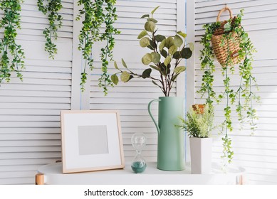 Photoframe, Vase, Hourglass, Plants And Various Decorative Objects On A Table In A Brightly Lit Room In Summer