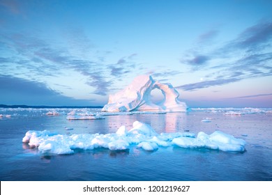 Photogenic and intricate iceberg with a hole under an interesting and colorful sky during sunrise with full moon. Disko bay, Greenland.