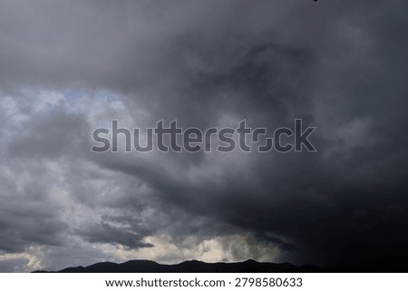 The photograph captures a dramatic sky filled with heavy, turbulent storm clouds that seem to gather with powerful intensity. Thick masses of deep charcoal and black clouds dominate the upper portion of the scene, creating an almost overwhelming sense of weight and movement. Beneath them, layers of softer gray clouds swirl and fold into one another, adding texture and depth across the sky. A pale opening near the center allows muted light to filter through, producing a striking contrast between darkness and illumination. The light appears trapped behind the clouds, as if the sun is struggling to break through the storm. The atmosphere feels tense, mysterious, and emotionally charged, suggesting an approaching downpour or a moment just before thunder rolls across the landscape. Overall, the image conveys nature’s raw power and unpredictability while creating a moody, cinematic feeling filled with drama and anticipation. The layered formations resemble waves of smoke and shadow, curling through the sky and enhancing the scene's haunting visual impact even further.
