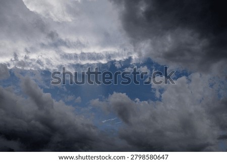 The photograph captures a dramatic sky filled with heavy, turbulent storm clouds that seem to gather with powerful intensity. Thick masses of deep charcoal and black clouds dominate the upper portion of the scene, creating an almost overwhelming sense of weight and movement. Beneath them, layers of softer gray clouds swirl and fold into one another, adding texture and depth across the sky. A pale opening near the center allows muted light to filter through, producing a striking contrast between darkness and illumination. The light appears trapped behind the clouds, as if the sun is struggling to break through the storm. The atmosphere feels tense, mysterious, and emotionally charged, suggesting an approaching downpour or a moment just before thunder rolls across the landscape. Overall, the image conveys nature’s raw power and unpredictability while creating a moody, cinematic feeling filled with drama and anticipation. The layered formations resemble waves of smoke and shadow, curling through the sky and enhancing the scene's haunting visual impact even further.
