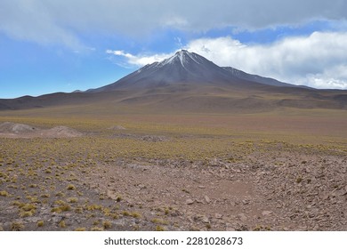 The photograph captures the grandeur of a volcano in the Atacama Desert Altiplano on a cloudy day. The majestic volcano is surrounded by a barren landscape with a few sparse plants, and the cloudy sky - Powered by Shutterstock