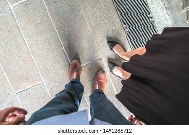 A Photograph Looking Down At A Caucasian Man And A Mixed Race African American Woman's Feet With Jeans And Dress Shoes And A Black Skirt Next To A Drainage Grate Outside On A Masonry Walkway.