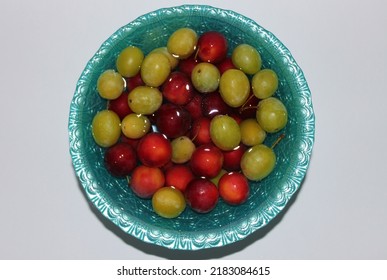 Photograph of luscious red plums and green plums on a white table, in water on a blue plate, fruits in water