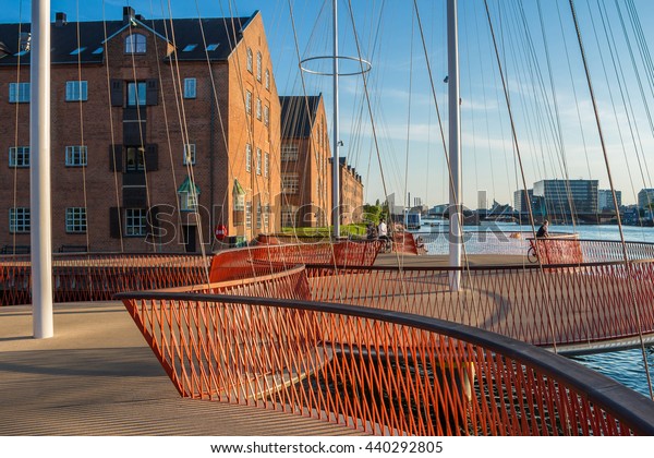Photograph of modern circle Bridge in\
Copenhagen, Denmark. Harbor with new circle bridge. Bridge with\
several circles, in shapes, form of masts. Red handrails and\
fences. Houses and sky on\
background.