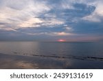 The photograph showcases a serene sunset descending over a calm seashore. The sun, a fiery orb, partially hidden by the cloud cover, casts a subtle reflection on the wet sand and gentle waves