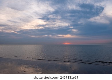 The photograph showcases a serene sunset descending over a calm seashore. The sun, a fiery orb, partially hidden by the cloud cover, casts a subtle reflection on the wet sand and gentle waves - Powered by Shutterstock