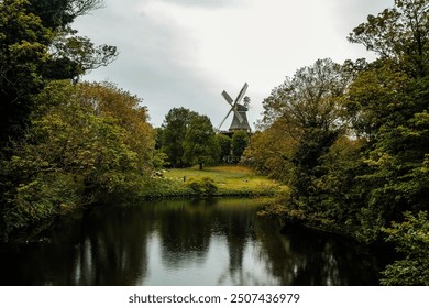 The photograph shows a forest with a traditional windmill standing on a grassy hill surrounded by lush trees. The tranquil pond reflects the greenery and overcast sky overhead. - Powered by Shutterstock