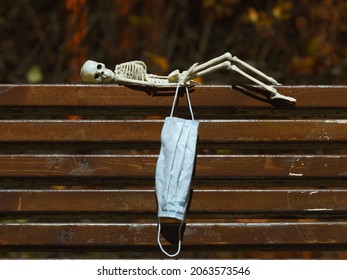 Photograph of a skeleton lies on a wooden bench at night. Tired abstract skeleton toy. He holding surgical mask. Natural dark background. Halloween concept.
