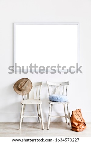 Photograph of a two vintage wooden chairs with a cushion, hat and handbag and an empty frame on the white wall