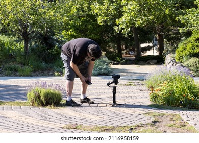 Photographer Bends Over A Camera Set Up On A Gimbal Recording Nature Scenes In A Garden