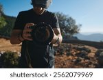 Photographer focuses his attention on nature scenes using a professional camera. The background shows a serene landscape under clear daylight, providing a peaceful and natural setting for photography.