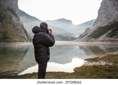 Photographer on assignment, holding a camera, taking photos of beautiful mountain landscape in the morning by a mountain lake with winter mist covered surface. 