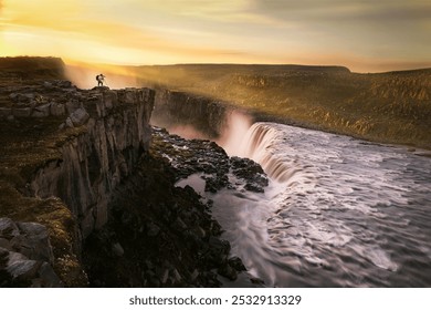 Photographer on rocky cliff captures a dramatic waterfall, with sunlight bathing the cascading water and misty atmosphere. - Powered by Shutterstock