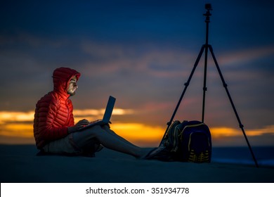 Photographer In Red Jacket With Hood Working With Laptop And Tripod On The Sand Dunes In The Early Morning. Time Lapse Filming Process