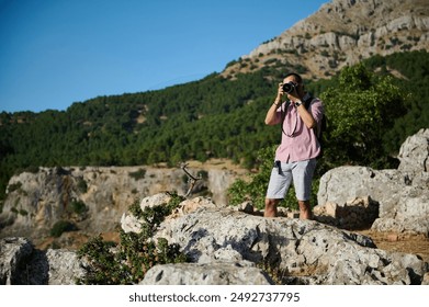 Photographer standing on rocky terrain capturing the scenic mountain landscape with a camera. Sunny day with clear blue sky and lush greenery in the background. - Powered by Shutterstock