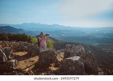 A photographer stands among rocky terrain, capturing the scenic mountain landscape on a bright sunny day. The horizon is filled with blue mountains and lush greenery under a clear sky. - Powered by Shutterstock