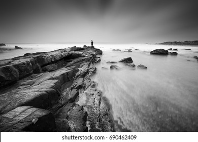 A photographer at the tip of a big rock watching big waves and being photographed at Karang Teraje, Sawarna Beach, Bayah, Banten, West Java Indonesia on long exposure. Black and white version
