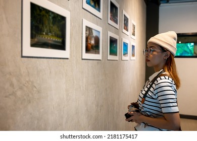 Photographer Visit At Photo Frame To Leaning Against At Show Exhibit Artwork Gallery, Asian Woman Hold Camera At Picture Art Gallery Collection In Front Framed Paintings Pictures On Wall And Looking