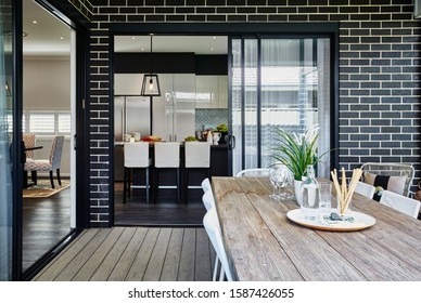 Photography Of Al Fresco Setting At Rear Of Modern Dark Brick Family Home With Outdoor Table Furniture Setting On Decking Area With View To Inside Of Kitchen Area With Breakfast Bar