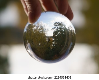 Photography Of Clear Transparent Crystal Ball. Human Fingers Holding It. Beautiful Autumn Is Coming. Green, Yellow Folliage Of Trees, Blue Clear Sky, Lake. Miracle View. Concept Of The Beaty In Nature