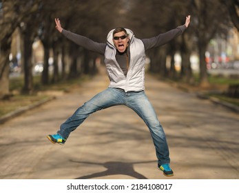 Photography Of Happy Handsome Man At Sunny Spring Day On The City Street. His Mood Is Good, Even Excellent. He Is Jumping Activelly. Stormy Joy. He Put On Blue Jeans And Insulated Vest. Close Up Image