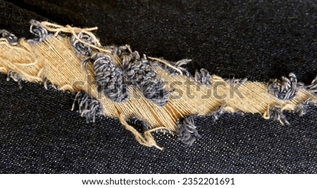 a photography of a piece of denim with a torn off piece of fabric, woollen fabric with fray edges and holes on a black background.