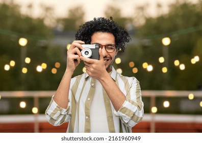 Photography, Profession And People And Concept - Happy Smiling Man Or Photographer In Glasses With Film Camera Over Party Lights On Roof Top Background