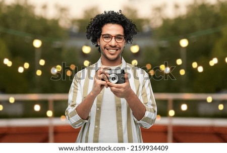 photography, profession and people and concept - happy smiling man or photographer in glasses with film camera over party lights on roof top background