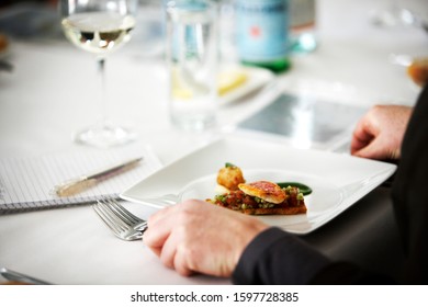 Photography of a table place settings in a fine dining restaurant with male food critic sitting at a table with a note pad and a fish and vegetable dish