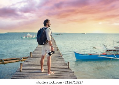 Photography and travel. Young man with rucksack holding camera standing on wooden fishing pier enjoying beautiful tropical sea view.