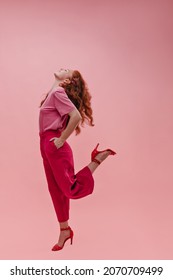 Photos of profile energetic caucasian young teenager with head thrown back and bent knee in studio. Curly-haired redhead girl holding hands in pockets dressed in casual clothes and high-heeled shoes.