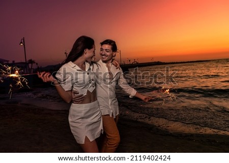 Photoshoot of young helthy beautiful caucasian couple, boy and girl lovers , dating at the beach - Man and woman celebrate New Year's Eve in holidays near the ocean with fireworks