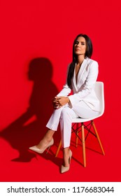 Photoshooting Studio Concept. Vertical Portrait Of Proud Arrogant Woman Sitting On Chair Leg By Foot Looking At Camera Isolated On Vivid Red Background