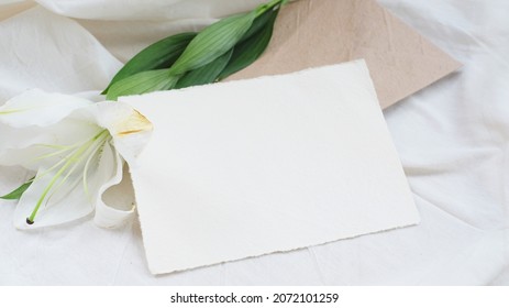 Photostock wedding styled composition. Feminine mockup scene with leaves, lily flower, brown recycled paper envelope, blank greeting card on white textured fabric background.
