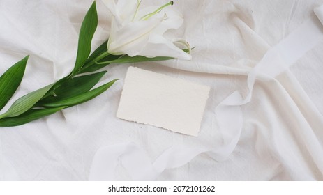 Photostock wedding styled composition. Feminine mockup scene with leaves, lily flower, blank greeting card recycled paper on white textured fabric background.