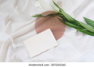 Photostock wedding styled composition. Feminine mockup scene with leaves, lily flower, blank greeting card recycled paper, round acrylic on white textured fabric background.