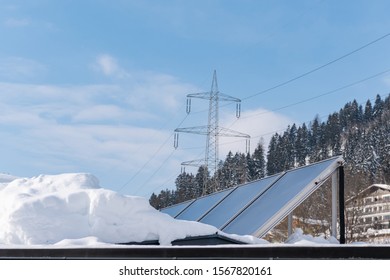 Photovoltaic Panels Partly Covered With Snow, Installed On The Roof Of The House. In The Background An Element Of The Power Grid - A High Voltage Power Pole With Overhead Wires.