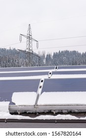 Photovoltaic Panels Partly Covered With Snow, Installed On The Roof Of The House. In The Background An Element Of The Power Grid - A High Voltage Power Pole With Overhead Wires.