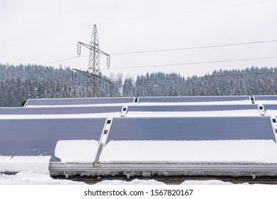 Photovoltaic Panels Partly Covered With Snow, Installed On The Roof Of The House. In The Background An Element Of The Power Grid - A High Voltage Power Pole With Overhead Wires.