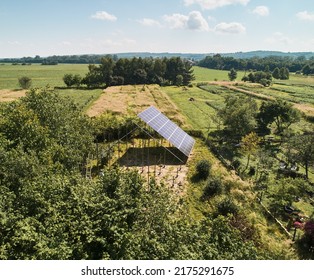 Photovoltaic Solar Panel And Supporting Structures For Solar Modules In Field With Green Tress. Grassy Area With Solar Panel System For Generating Electricity Through Photovoltaic Effect.