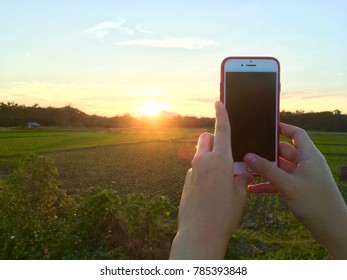 PHRAE, THAILAND - JAN 2, 2018 : Woman's Hand Holding The Phone And Take A Photo Of The Sun And Mountain In Thailand By The Camera App On An Apple IPhone 7 Red.