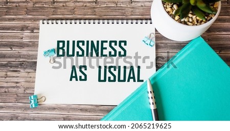 The phrase Business as usual on a notepad and a diary with a pen on a wooden table.