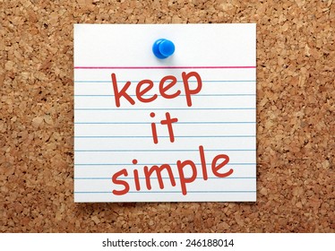 The phrase Keep It Simple in red ink on a white index card pinned to a cork notice board as a reminder
