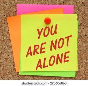 The phrase You Are Not Alone in red text on a yellow sticky note pinned to a cork notice board as a reminder
