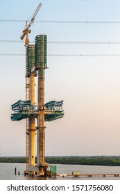 Phuoc Khanh, Vietnam - March 13, 2019: Long Tau River At Sunset. Closeup Of Western Pylon Of Phuoc Khanh Suspension Bridge Under Construction. Gray Water, Green Belt And Gray Sky. Sunset Colors Concre