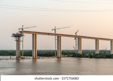 Phuoc Khanh, Vietnam - March 13, 2019: Long Tau River At Sunset. Western On-ramp Of Phuoc Khanh Suspension Bridge Under Construction. Gray Water, Green Belt And Gray Sky. Sunset Colors Concrete Brown.