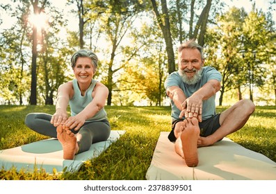 Physical activity outside in summer. Portrait of aged couple sitting on yoga rugs and doing stretching exercises in park. Cheerful man and woman looking at camera while reaching for toes and smiling.