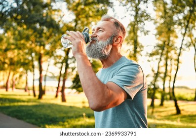 Physical activity in park. Side view of thirsty aged man drinking fresh water from fitness bottle after intense workout on fresh air. Athletic male maintaining water balance during outdoor training. - Powered by Shutterstock